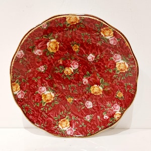 May include: A round, red ceramic plate with a scalloped edge and a gold rim. The plate is decorated with a pattern of yellow, pink, and white roses and green leaves.