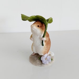 May include: A detailed figurine of a hamster sheltering under a green leaf. The hamster has brown and white fur and stands on a small, grey stone base with purple and white flowers. A decorative ornament.