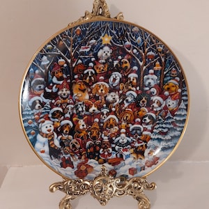 May include: A round, white porcelain plate with a gold rim and a festive Christmas scene featuring a large group of dogs wearing Santa hats and other holiday attire. The plate is on a gold metal stand.