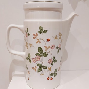 May include: White ceramic jug with a lid, handle, and spout. The jug is decorated with a pattern of pink strawberry blossoms, green leaves, and red strawberries. The jug is on a white surface.