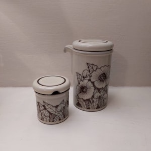 May include: A set of two white ceramic condiment containers with brown floral designs. The larger container has a pour spout and a lid. The smaller container has a lid.