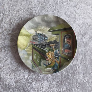 May include: A green and blue train engine with a steam plume and a frog peeking out of the cab window. The train is on a track with a green hillside in the background. The words "G.E.R." are visible on the side of the train.