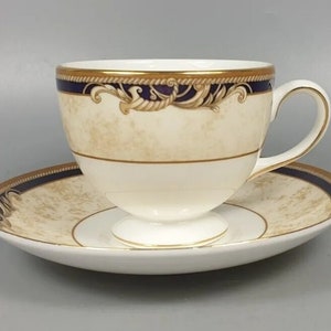May include: A cream-coloured teacup and saucer set. The cup and saucer have a gold rim and a navy blue band with a decorative gold design. The cup sits on a small pedestal base. The saucer is round.