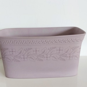 May include: A rectangular, lavender-coloured planter with a floral design. The planter has a decorative border near the top and a detailed floral pattern around the middle. The planter is designed for indoor or outdoor use.