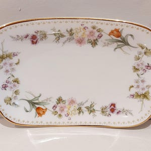 May include: An oval porcelain serving tray with a white base and a gold-trimmed edge. The tray features a delicate floral pattern around the perimeter, showcasing various flowers in shades of pink, yellow, orange, and green foliage.