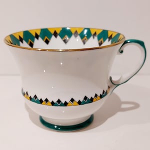 May include: White teacup with a green handle and base, and a gold rim. The cup features a geometric pattern of black diamonds, yellow triangles, and green accents around the rim and base. A classic tea cup.