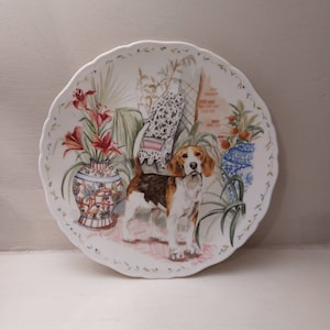 May include: A white ceramic plate with a scalloped edge features a colourful illustration of a beagle dog standing in a garden setting. The dog is brown and white with a black nose and brown eyes. The garden is filled with green plants, red flowers, and a blue and white vase.
