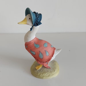 May include: A ceramic duck figurine wearing a blue bonnet and a red shawl with blue teardrop patterns. The duck has a yellow beak and feet, and stands on a small, textured base.
