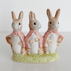 May include: A ceramic ornament depicting three rabbits wearing pink capes, standing on a green grassy base. The rabbits have brown ears and eyes, and are holding baskets. The ornament is a decorative item.