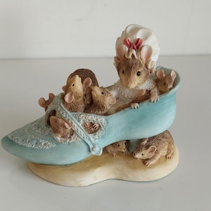 May include: A charming figurine depicting a blue shoe adorned with lace, housing several brown mice. The shoe rests on a beige base. One mouse is wearing a white bonnet with a red bow.