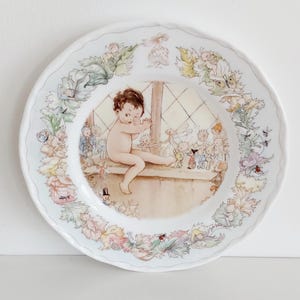 May include: A white decorative plate with a scalloped edge. The plate features a whimsical illustration of a child and fairies near a window. The border is adorned with floral and leaf motifs in pastel colours, with small insects.