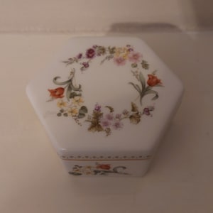 May include: A white hexagonal ceramic trinket box with a floral design. The design features a wreath of pink, purple, yellow, and orange flowers with green leaves.