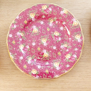 May include: A round, claret-coloured plate featuring a floral pattern of yellow and pink roses. The plate has a scalloped edge and a gold rim. The floral design covers the entire surface of the plate.