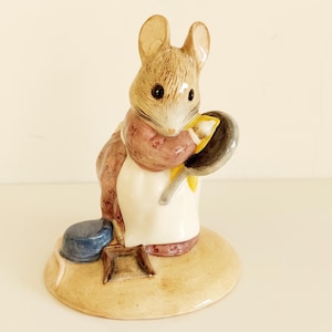 May include: A ceramic mouse figurine dressed as a chef, holding a frying pan. The mouse has a light brown coat, a pink scarf, and a white apron. A blue pot and small wooden stool are at its feet. The figurine is on a tan base.