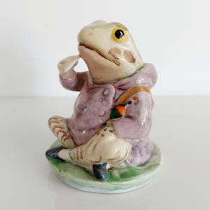 May include: A ceramic frog figurine, wearing a lavender coat and holding a small object. The frog has yellow eyes and sits on a green base. The figurine has a vintage aesthetic, reminiscent of a storybook character.