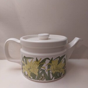 May include: White ceramic teapot with a floral design. The teapot has a lid and a handle. The flowers are yellow and green.