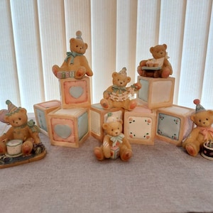 May include: A set of seven teddy bear figurines wearing party hats and holding birthday cake and presents. The bears are arranged on top of stacked blocks with a pastel colour scheme and floral designs.