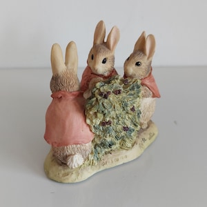 May include: A decorative figurine of three rabbits in pink capes, gathered around a bush with green leaves and purple berries. The rabbits have tan fur and are on a cream-coloured base.