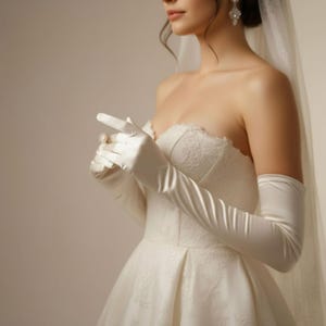 May include: A bride wearing a white satin wedding dress and long, elegant white satin gloves. The gloves extend past the elbows. The dress has a lace bodice and a full skirt.
