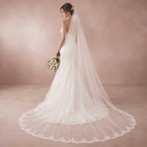 May include: A white floor-length wedding dress with a fitted bodice and a flared skirt featuring lace detailing. A long, sheer, floor-sweeping veil with a scalloped lace border is attached to the hair, cascading down the back of the dress.