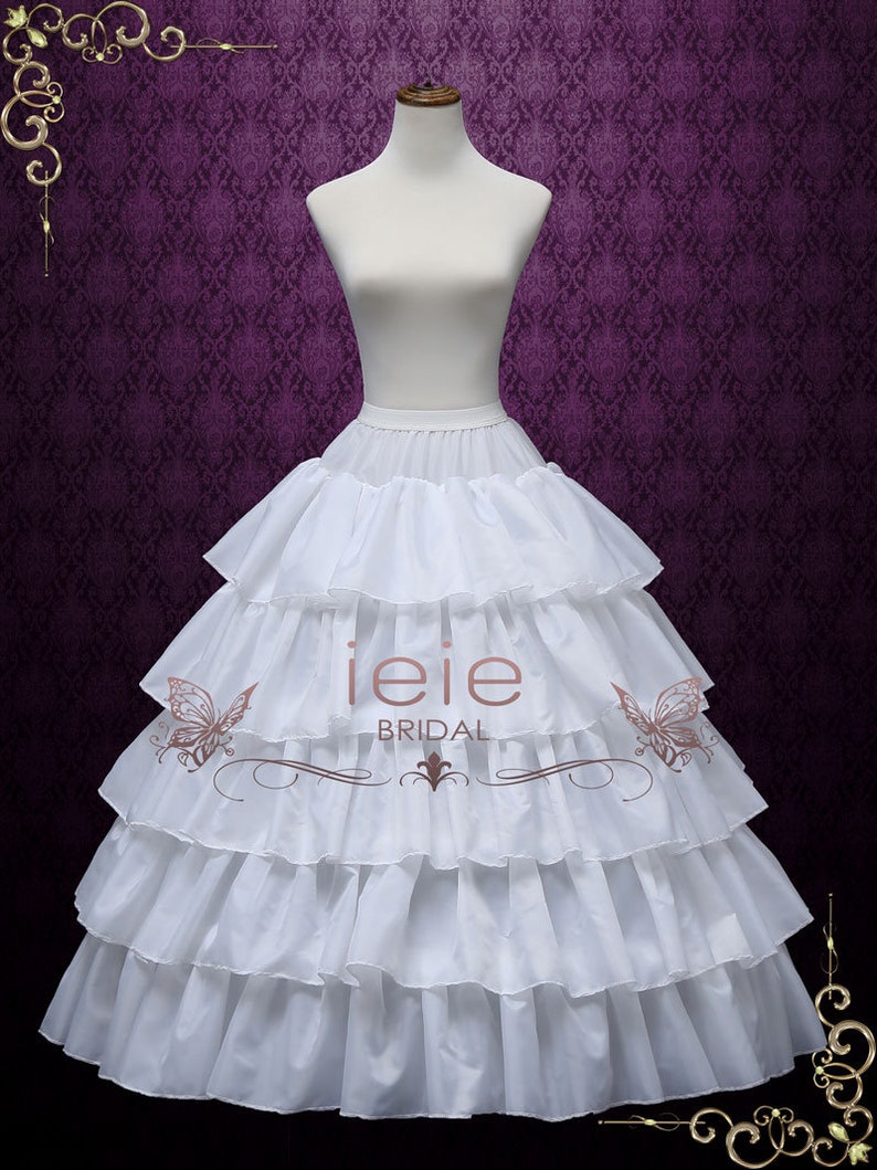 Puede incluir: Un enaguas blanco de varias capas con un borde con volantes. El enaguas est&aacute; en un maniqu&iacute; blanco sobre un fondo morado. El texto "ieie BRIDAL" es visible en el enaguas.