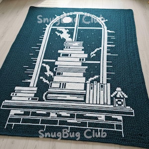 Puede incluir: Una alfombra de ganchillo de color verde azulado con un gráfico blanco de una ventana con una pila de libros, una estantería y una casita. El texto "SnugBug Club" es visible. La alfombra es rectangular y parece hecha a mano.