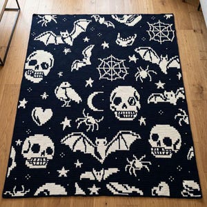 May include: A rectangular crochet blanket featuring a dark navy blue background with a repeating white pixelated pattern of skulls, bats, spiderwebs, spiders, stars, a crescent moon, a heart, and a bird, all arranged in a dense, scattered layout.