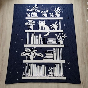 Könnte beinhalten: Eine marineblaue Häkeldecke mit einem weißen Bücherregal-Design. Das Design umfasst Regale mit Büchern, Topfpflanzen und einer Katze. Die Decke hat ein strukturiertes Aussehen und ist wahrscheinlich handgefertigt.