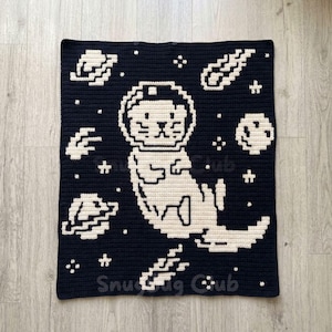 Pode incluir: Um cobertor de crochê azul marinho e creme com uma lontra de desenho animado em um capacete espacial flutuando entre planetas, estrelas e estrelas cadentes. As palavras "Snug Club" estão na parte inferior.