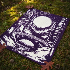 Puede incluir: Una manta de ganchillo morada con un diseño de bosque blanco. El diseño presenta una luna llena, árboles y un puente. La manta es rectangular y tiene el texto "Snug Club" en la esquina superior derecha.