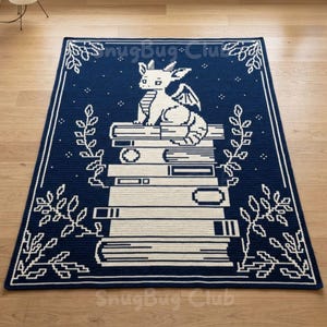 Può includere: Una coperta all'uncinetto blu navy e crema con un drago seduto su una pila di libri. La coperta ha un bordo bianco con decorazioni floreali e il testo "SnugBug Club".