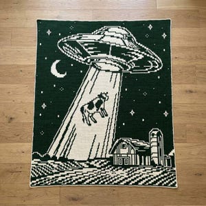 Könnte beinhalten: Eine rechteckige Häkeldecke mit dunkelgrünem Hintergrund und einem weißen, pixeligen Design eines UFOs, das über einem Bauernhof schwebt. Eine Kuh wird von einem Lichtstrahl des Raumschiffs entführt, ergänzt durch eine Mondsichel und Sterne.