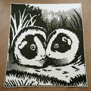 Pode incluir: Um cobertor de crochê em preto e branco com dois texugos estilizados em um cenário de floresta. O cobertor tem um design pixelado, com os texugos voltados um para o outro. O cobertor é retangular e parece ser feito à mão.