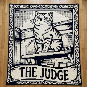 Könnte beinhalten: Eine rechteckige Häkeldecke in Dunkelblau und Creme. Das Design zeigt eine Katze, die auf einem Sims innerhalb eines dekorativen Rahmens sitzt, mit dem Text THE JUDGE in fetten Großbuchstaben am unteren Rand der Decke.