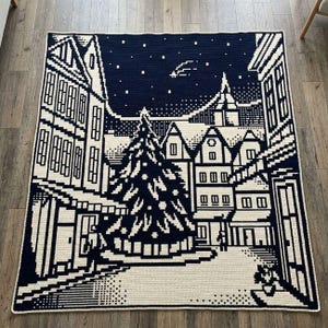 May include: A rectangular crochet blanket featuring a dark navy blue and cream pixelated winter scene. The design depicts a town square with a large central Christmas tree, surrounding multi-story buildings, and a starry night sky with a comet.