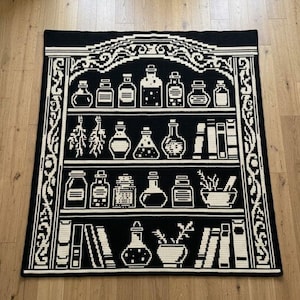 May include: A rectangular crochet blanket featuring a black and white pixelated design of a bookshelf. The shelves are filled with various glass potion bottles, jars, books, and bundles of dried herbs, all framed by an ornate, decorative border.
