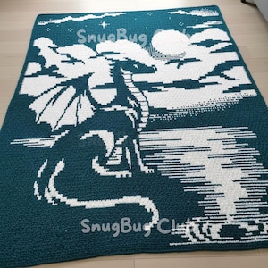 May include: A teal and white crocheted blanket with a dragon motif. The white dragon has detailed wings and tail, set against a night sky with a full moon and clouds. The text "SnugBug" is visible.
