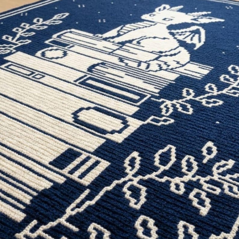 Pode incluir: Um cobertor de malha azul marinho e creme com um desenho de drag&atilde;o. O cobertor mostra um drag&atilde;o sentado em livros, com elementos florais decorativos. O design &eacute; detalhado.