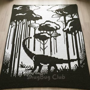 Könnte beinhalten: Eine schwarz-weiß gehäkelte Decke mit einem Dinosaurier in einer Waldszene. Die Decke hat einen dunkelgrünen Rand und die Worte "SnugBug Club" sind sichtbar. Das Design ist pixelig.
