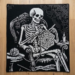 Op de afbeelding: Een zwart-witte gehaakte deken met een grafisch ontwerp van een skelet dat in een stoel zit en een zeshoekig stuk breit. Een bol wol ligt op de armleuning van de stoel en een brandende kaars staat op een tafeltje ernaast.