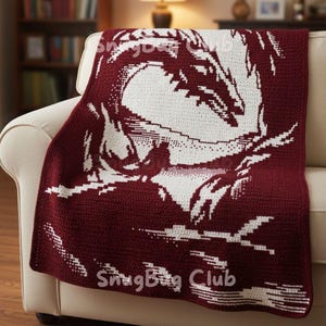 May include: A burgundy and white crocheted blanket featuring a dragon design. The dragon is white against a burgundy background. The blanket is draped over a beige sofa. The text "SnugBug Club" is visible.