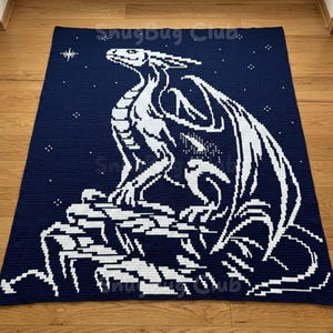 以下が含まれることがあります： ネイビーブルーと白の、ドラゴンのデザインの編み物ブランケット。白いドラゴンは白い岩の上にいます。背景はネイビーブルーで、白い星があります。「SnugBug Club」の文字が見えます。