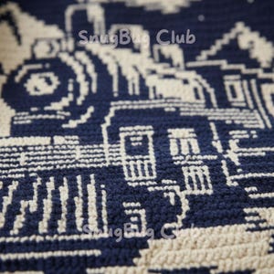 Op de afbeelding: Close-up van een marineblauwe en cr&egrave;mekleurige gehaakte deken. De deken heeft een herhalend patroon van gebouwen en de tekst "SnugBug Club". Het ontwerp is een gestructureerd, gedetailleerd patroon.