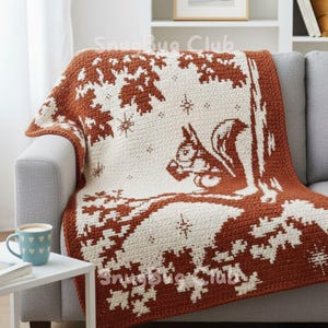 May include: A crocheted blanket in shades of cream and rust brown, featuring a squirrel design. The blanket is draped over a gray sofa. The design includes a squirrel, trees, and snowflakes. The blanket is labeled "SnugBug Club".