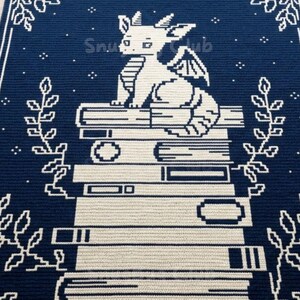 Pode incluir: Um tapete de croch&ecirc; azul marinho e branco com um drag&atilde;o sentado em cima de uma pilha de livros. O tapete tem uma borda com padr&otilde;es florais e de estrelas, e o texto "Snu Club" &eacute; vis&iacute;vel.