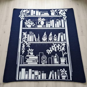 May include: A navy blue and white crocheted blanket featuring a bookshelf design. The design includes stacks of books, potted plants, and decorative objects. The blanket is rectangular and appears to be made of soft yarn.