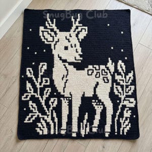Könnte beinhalten: Eine quadratische, dunkelmarineblaue Häkeldecke mit einem weißen Hirschdesign. Der Hirsch ist von weißem Laub und kleinen weißen Sternen umgeben. Der Text "SnugBy Club" befindet sich in den oberen Ecken und in der unteren rechten Ecke.