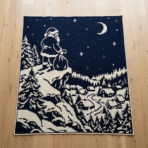 Könnte beinhalten: Eine rechteckige Häkeldecke in Dunkelblau und Creme. Das Design zeigt die Silhouette des Weihnachtsmanns, der auf einer verschneiten Klippe über einem Dorf steht, mit einer Mondsichel und Sternen am nächtlichen Himmel.