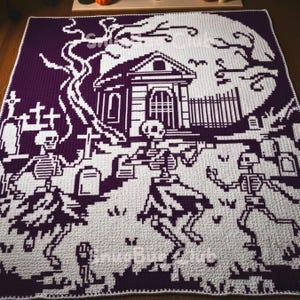 May include: A crocheted blanket featuring a Halloween-themed design. The blanket showcases a purple and white color scheme, with a graveyard scene, including skeletons, a haunted house, and a full moon. The blanket is rectangular.