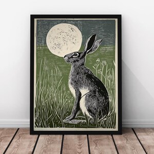 May include: A framed art print depicting a hare in a field, looking up at a full moon. The artwork is in black and white, set against a green and dark blue backdrop. The print is framed in black.
